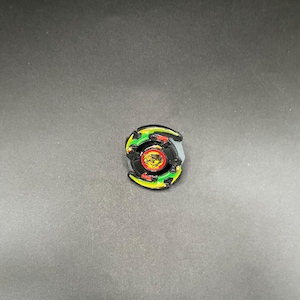 May include: A small, colorful toy with a circular design. The toy features a black center with a yellow and red emblem, surrounded by green, yellow, and red accents. The outer edges are gray. The toy is set against a dark background.