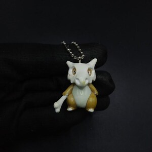 Puede incluir: Un pequeño llavero con figura de Cubone. La figura es principalmente marrón y blanca, con un hueso en la mano. El llavero está sujeto a una cadena plateada. El fondo es de color oscuro.