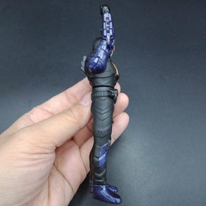 May include: A side view of a black action figure with purple and orange accents. The figure is holding a purple weapon. The figure is in a standing position. The figure is held by a hand.