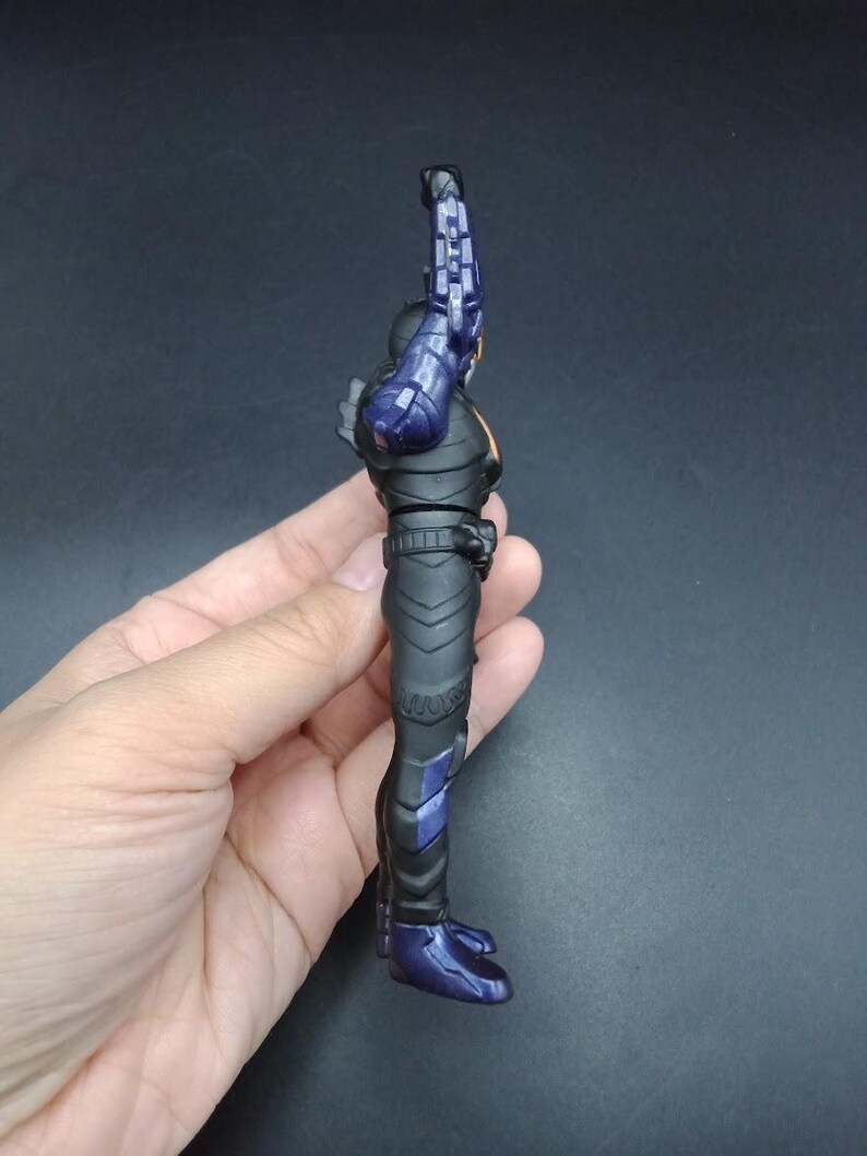 May include: A side view of a black action figure with purple and orange accents. The figure is holding a purple weapon. The figure is in a standing position. The figure is held by a hand.