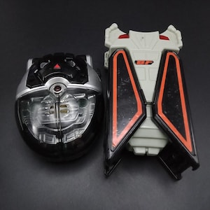 May include: Two futuristic toy devices. One is a black and silver oval with a clear center, the other is a white and black rectangular device with orange accents. The devices are on a dark surface.