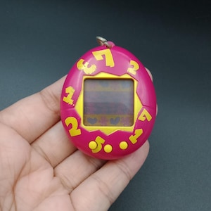 May include: A pink handheld digital pet game with yellow numbers and buttons. The screen displays a pixelated graphic. The device has a silver metal loop at the top for attaching to a keychain. The device is held in a hand.
