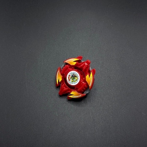 May include: Red and yellow plastic spinning top with a black and white design in the center. The top has the words "Attack" and "Balance" printed on it.