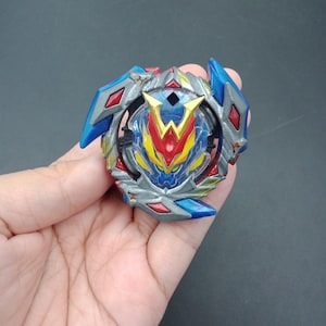 Op de afbeelding: Een veelkleurige speelgoedtol in blauw, rood, geel en zilver. Het speelgoed heeft een centraal embleem en geometrische componenten. De tol wordt in een hand gehouden tegen een donkere achtergrond.