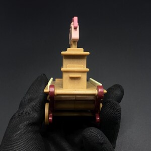 May include: A small, tan-colored toy wagon with red wheels and accents. The wagon has a rectangular body with a tall, pink structure on top. The toy is held in a black gloved hand.