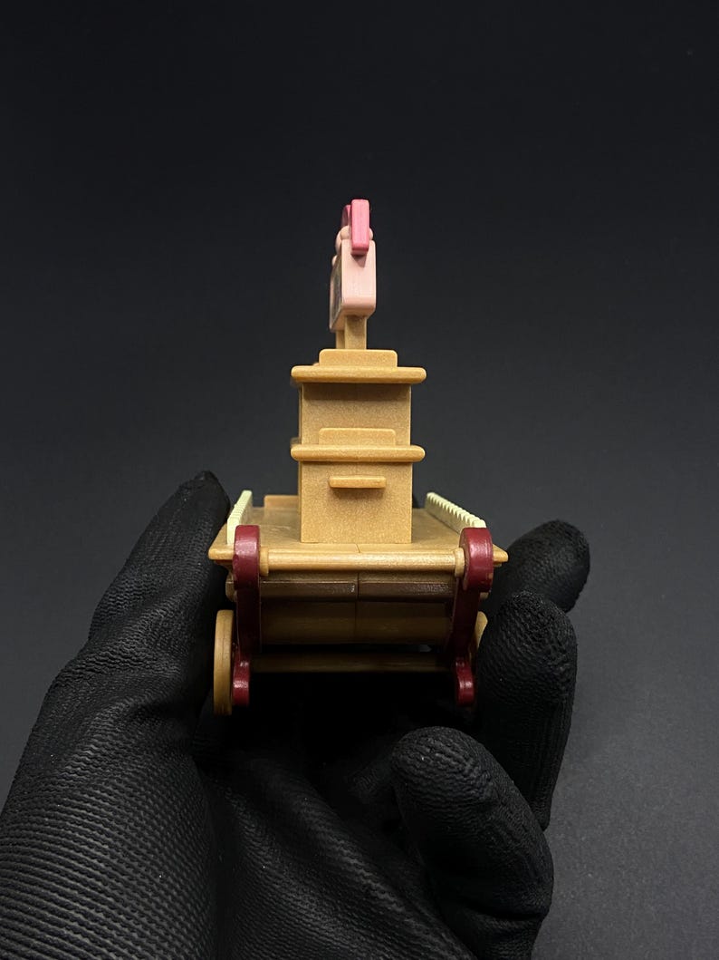 May include: A small, tan-colored toy wagon with red wheels and accents. The wagon has a rectangular body with a tall, pink structure on top. The toy is held in a black gloved hand.