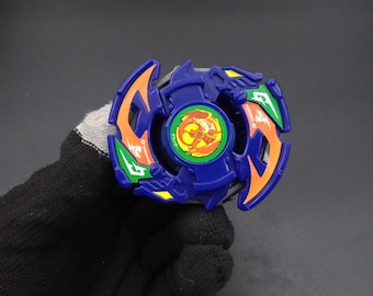 May include: A blue, orange, and green spinning top toy. The toy has a circular design with a central emblem and curved blades. The colors are vibrant, and the toy appears to be made of plastic. The toy is held by a gloved hand.