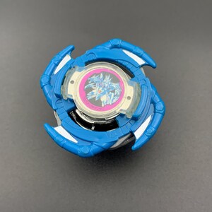 Puede incluir: Un juguete de peonza azul y plateado con un emblema central circular con un borde rosa y una ilustración gráfica de una criatura blanca. El juguete tiene cuatro brazos azules curvos en forma de garra con detalles plateados.