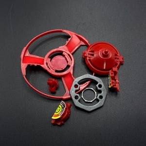May include: A red and gray Beyblade toy with a spinning top, a metal frame, and a red plastic piece with the word "Nitro" on it.