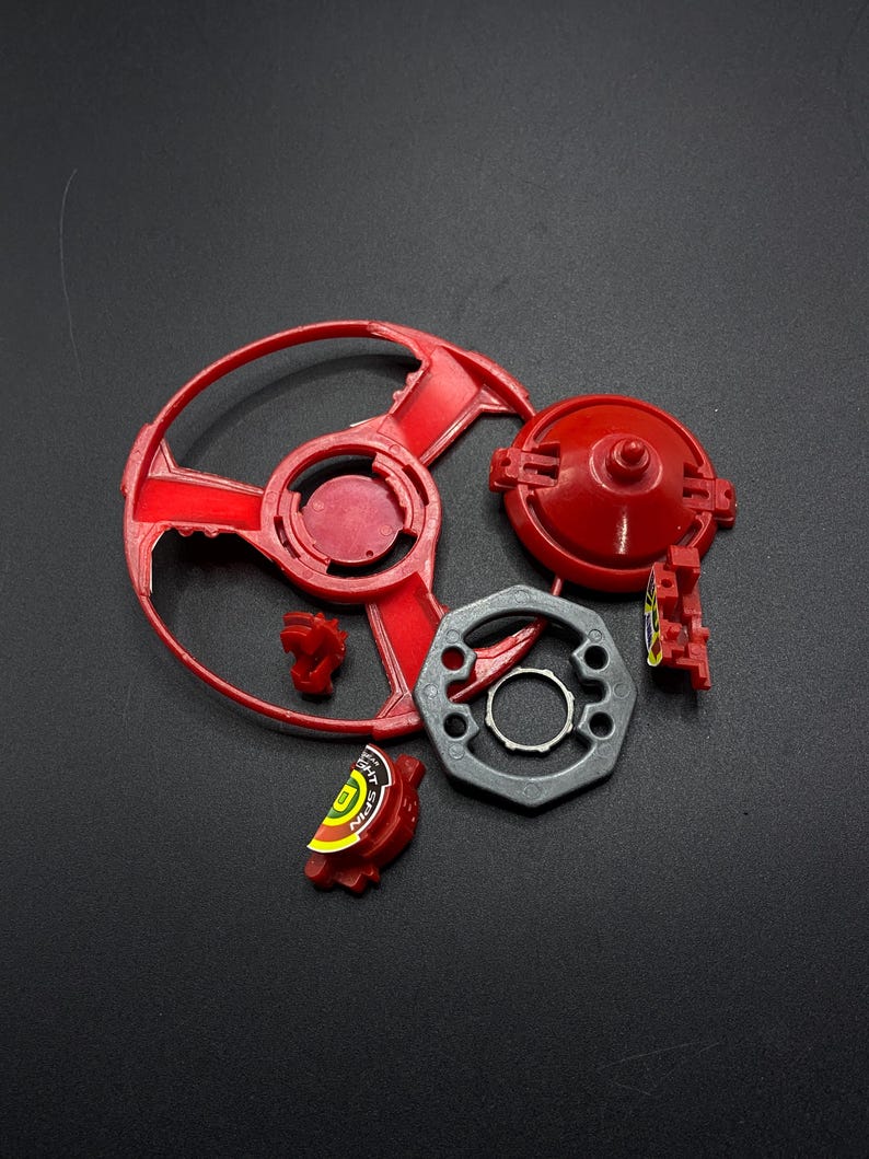 May include: A red and gray Beyblade toy with a spinning top, a metal frame, and a red plastic piece with the word "Nitro" on it.