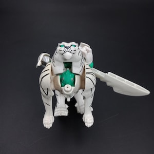 May include: A white and black tiger-shaped toy with green accents and a white blade. The toy is in a crouching position, with its mouth open. The toy is on a black background.