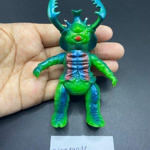 May include: A green and blue toy figure with large, curved horns and a red mouth. The figure has a segmented torso with red and blue accents. The figure is held in a hand, and a white label with the text "B627 Pb246" is visible.