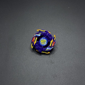 Könnte beinhalten: Ein blaues, gelbes und rotes Kreiselspielzeug. Das Spielzeug hat ein kreisförmiges Design mit mehreren Schichten und Details. Die Mitte ist blau mit einem gelben Emblem, und die äußeren Ränder sind gelb mit roten und blauen Akzenten.