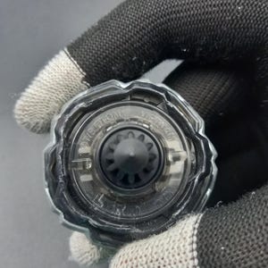 Pu&ograve; includere: Primo piano di un trottola giocattolo nera e trasparente. La parte superiore ha un design circolare con un ingranaggio nero centrale e un anello esterno trasparente. Il testo "TIS-GTON" e "VIETNAM" &egrave; visibile sulla parte superiore.