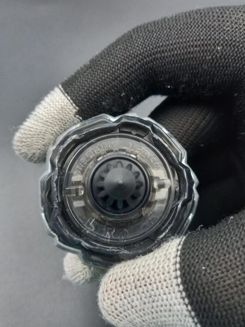 Pu&ograve; includere: Primo piano di un trottola giocattolo nera e trasparente. La parte superiore ha un design circolare con un ingranaggio nero centrale e un anello esterno trasparente. Il testo "TIS-GTON" e "VIETNAM" &egrave; visibile sulla parte superiore.