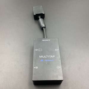 Peut inclure: Adaptateur MultiTap Sony noir pour PlayStation 2. L'adaptateur a une base rectangulaire avec le mot "MULTITAP" et le logo PlayStation 2. Il comporte quatre ports étiquetés A, B, C et D. Un câble s'étend du haut.