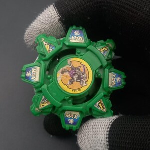 May include: A green and yellow toy with a circular design. The toy has a central image of a purple and yellow creature and the word "MAX" on several sections. The toy is held by a person wearing a black glove.