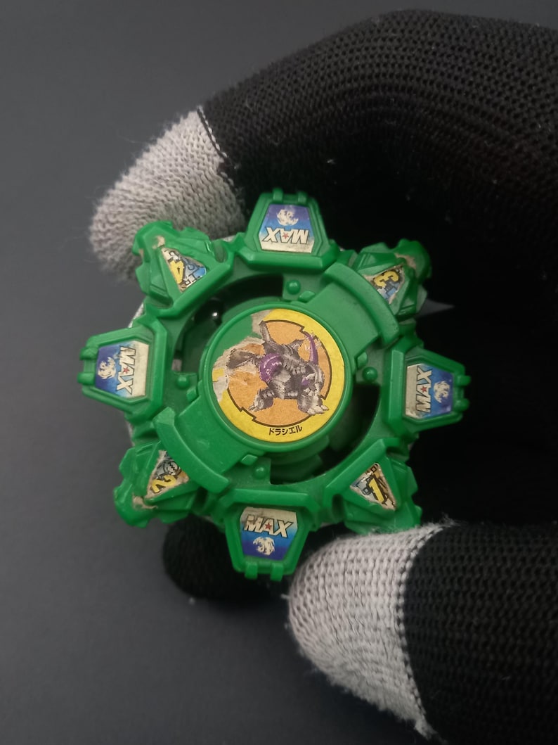 May include: A green and yellow toy with a circular design. The toy has a central image of a purple and yellow creature and the word "MAX" on several sections. The toy is held by a person wearing a black glove.