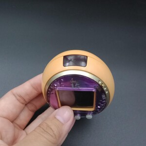 May include: A handheld, retro-style electronic game with a rounded, peach-colored body. It features a purple screen displaying the number "15", a gold-colored trim, and two gray buttons. The device is held against a dark background.