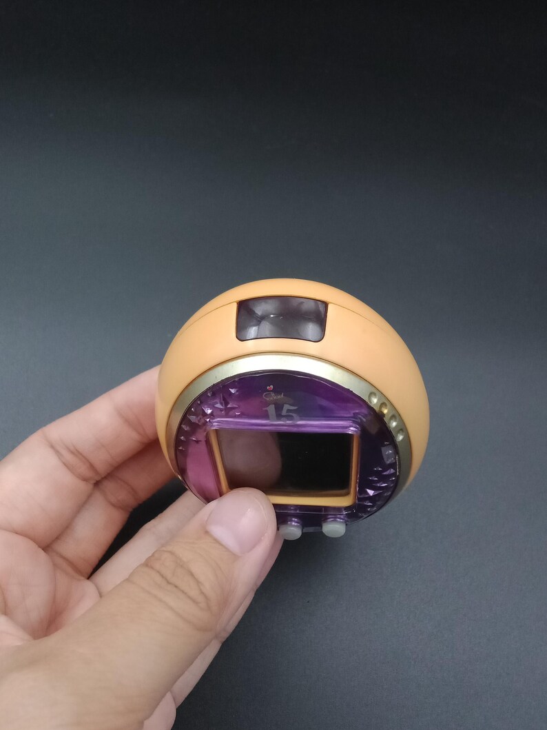 May include: A handheld, retro-style electronic game with a rounded, peach-colored body. It features a purple screen displaying the number "15", a gold-colored trim, and two gray buttons. The device is held against a dark background.