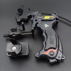 May include: A black and grey Beyblade launcher set. The set includes a launcher grip with "EZ GRIP" text, a launcher, and a separate winder. The launcher grip has a red accent and a bird emblem.