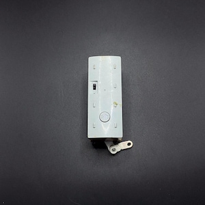 Puede incluir: Un objeto rectangular gris claro con una pequeña pieza blanca con bisagras adjunta. El objeto tiene varias pequeñas aberturas cuadradas y un botón circular. El fondo es gris oscuro sólido.