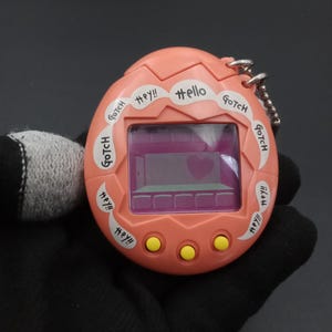 Puede incluir: Un dispositivo digital portátil de color coral con una pantalla que muestra un corazón. El dispositivo tiene las palabras "Hello", "Hey!!" y "Gotch" impresas. Tiene tres botones amarillos y una cadena plateada.