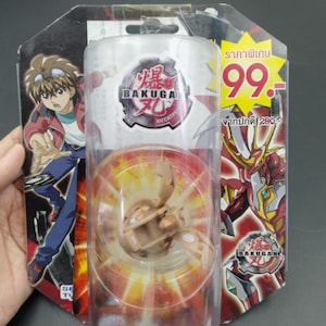 Op de afbeelding: Een bruin Bakugan Mechtainer-speelgoed in een doorzichtige plastic verpakking. De verpakking bevat een cartoonillustratie van een personage en het Bakugan-logo. De verpakking bevat ook tekst in een vreemde taal.