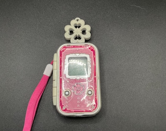 May include: A pink and silver handheld electronic device with a small screen and a pink strap. The device has a clover-shaped charm at the top and two circular buttons on the front. The screen is blank.