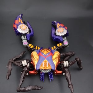 May include: A multi-colored crab-like toy with articulated legs and claws. The toy features a gold, red, purple, and black color scheme. The claws are purple with orange and silver accents. The body has a gold and red finish.
