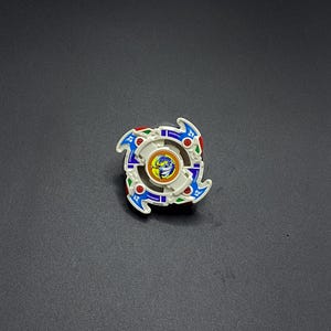 Könnte beinhalten: Ein weißer, blauer, roter und grüner Beyblade aus Plastik mit einem Drachenmotiv in der Mitte. Der Beyblade hat eine vierzackige Sternform.