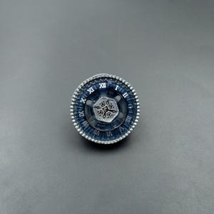 Pode incluir: Um brinquedo de pião circular com um anel exterior azul escuro e algarismos romanos brancos de I a XII. O componente central é um hexágono metálico prateado com um design gravado simétrico e intrincado e um pequeno recorte circular central.
