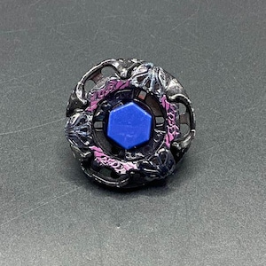 May include: A black and purple spinning top toy with a blue center. The top has a circular design with intricate details and a hexagonal blue piece in the center. The toy is designed for spinning and battling.