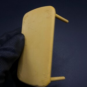 May include: A light yellow, rectangular object with two cylindrical pegs extending from the side. The object is held by a gloved hand against a dark blue background. The object has some minor imperfections.