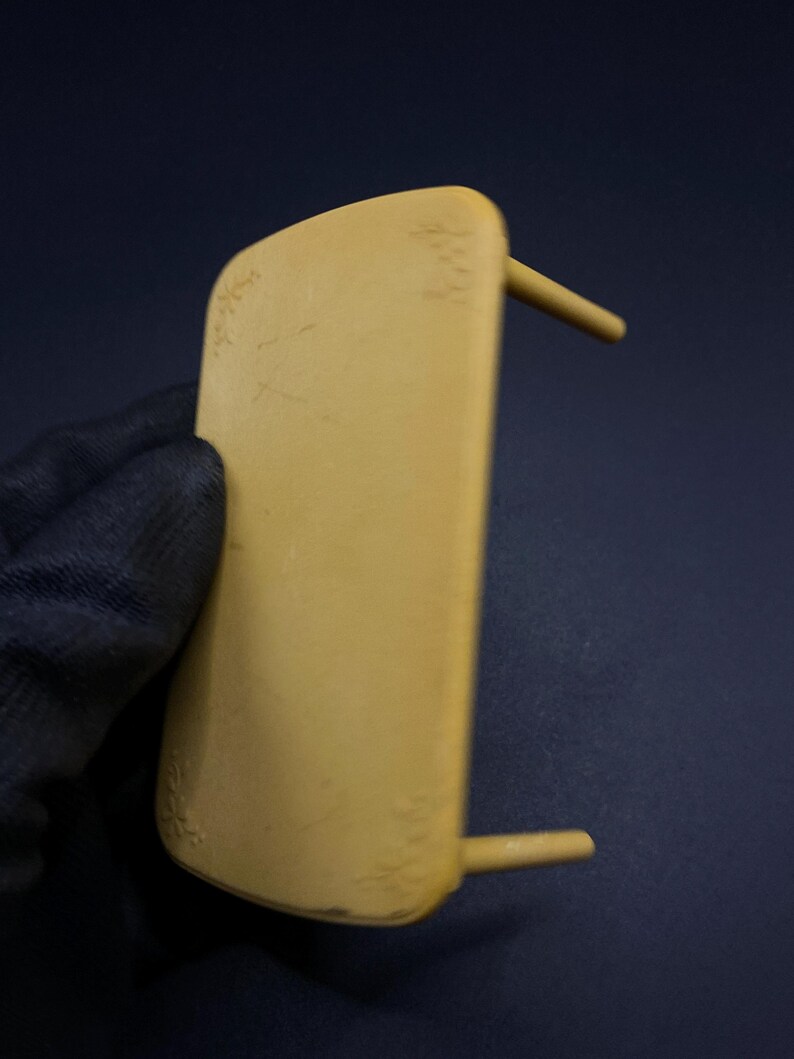 May include: A light yellow, rectangular object with two cylindrical pegs extending from the side. The object is held by a gloved hand against a dark blue background. The object has some minor imperfections.