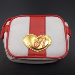 Pode incluir: Uma bolsa retangular branca e vermelha com um emblema dourado em forma de coração. A bolsa tem uma faixa vermelha no centro e acabamentos vermelhos. O emblema apresenta a letra "A" em uma fonte decorativa.