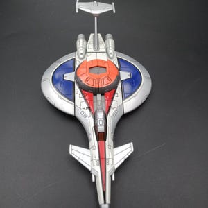 May include: A silver and red toy spaceship with blue and orange accents. The toy has a central body with wings and a vertical stabilizer. The design includes multiple engines and a cockpit. The spaceship is a futuristic design.