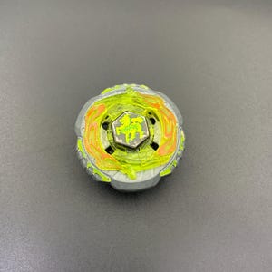 Puede incluir: Una peonza circular con una base de metal gris y un anillo de plástico amarillo neón translúcido. El centro incluye un emblema hexagonal con un gráfico de un lobo verde, rodeado de detalles en forma de llamas naranjas sobre el plástico amarillo.