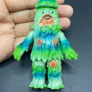 May include: A green and blue monster figurine with a mushroom-shaped head. The toy has a shaggy, textured appearance with red accents on the chest and legs. The figure has a wide mouth and small eyes.