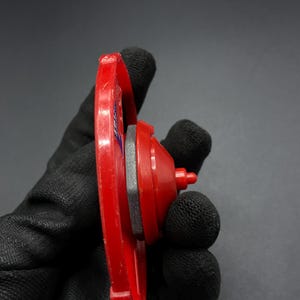 May include: A red and gray plastic toy top with a spinning disc and a pointed tip. The top is held in a black glove.