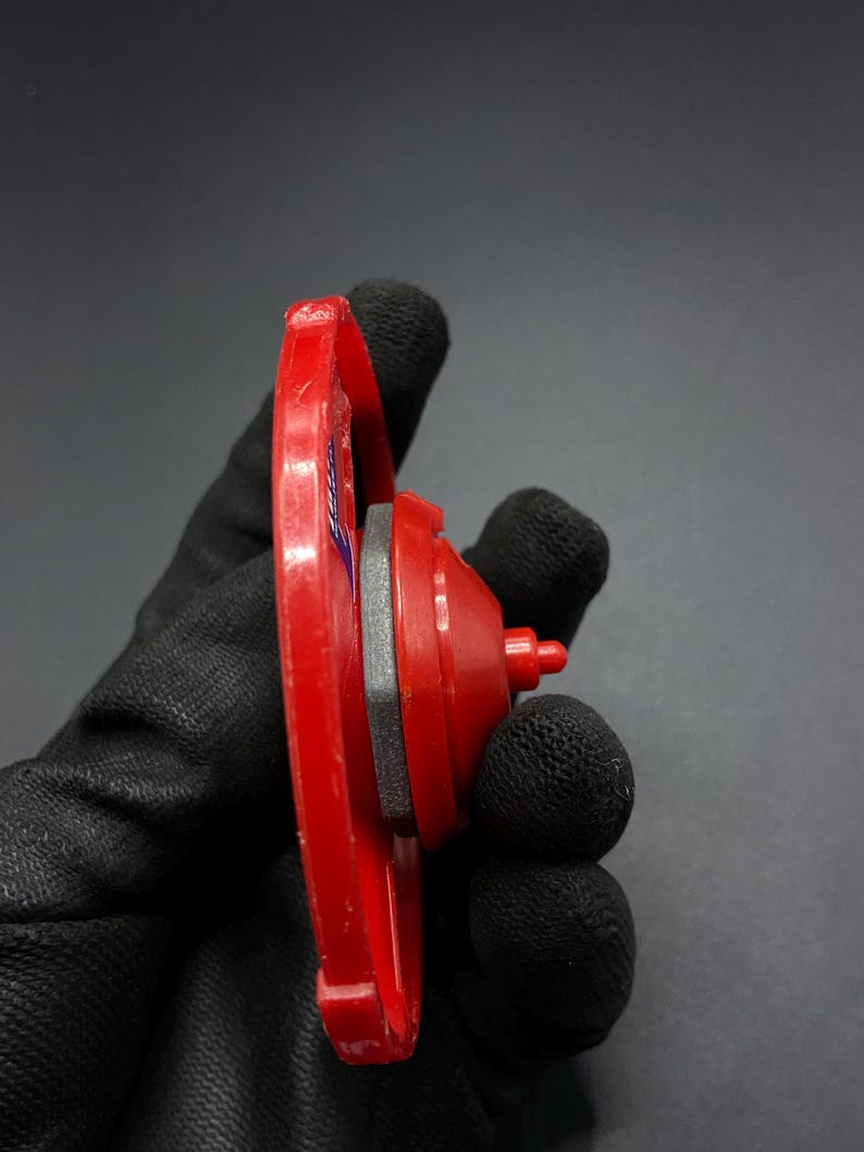 May include: A red and gray plastic toy top with a spinning disc and a pointed tip. The top is held in a black glove.
