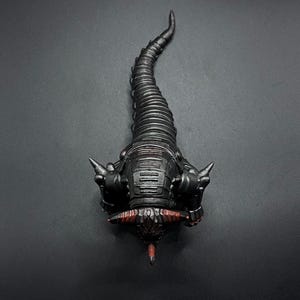 May include: A black and red detailed, articulated, segmented, mechanical creature with a long tail and sharp horns. The creature is posed in a curled position with its head facing the camera.