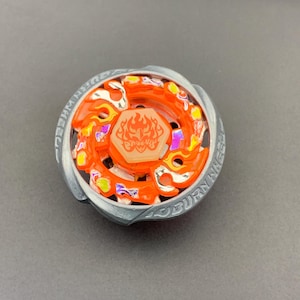 Könnte beinhalten: Ein kreisförmiges Kreiselspielzeug mit einem leuchtend orangefarbenen Mittelstück, das eine Flammengrafik und das Wort Phoenix zeigt. Es ist von einem metallisch silbernen Außenring umgeben, auf dem der Text Burn Wheel eingeprägt ist.