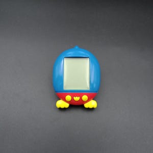 May include: A blue, red, and yellow handheld digital pet game shaped like a penguin. The device has a rectangular screen and yellow feet. The top of the device is blue, the middle is red, and the bottom has yellow feet.
