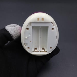 Puede incluir: Un dispositivo electr&oacute;nico blanco en forma de huevo con un compartimento de bater&iacute;a abierto. El dispositivo tiene una parte superior rosa y dos peque&ntilde;as aberturas rectangulares. El compartimento de la bater&iacute;a est&aacute; dise&ntilde;ado para contener dos bater&iacute;as. El dispositivo es sostenido por una mano enguantada.