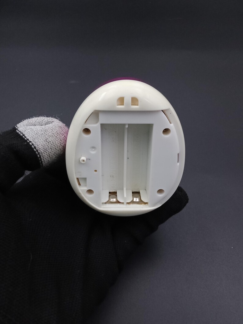 Puede incluir: Un dispositivo electr&oacute;nico blanco en forma de huevo con un compartimento de bater&iacute;a abierto. El dispositivo tiene una parte superior rosa y dos peque&ntilde;as aberturas rectangulares. El compartimento de la bater&iacute;a est&aacute; dise&ntilde;ado para contener dos bater&iacute;as. El dispositivo es sostenido por una mano enguantada.