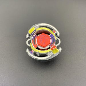 May include: A circular metal and plastic spinning top toy featuring a central orange hexagonal piece with a purple dragon-like graphic. The toy has silver curved outer edges, yellow accents, and purple patterned sections surrounding the centre.
