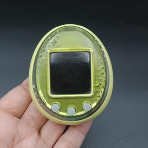 May include: A handheld, egg-shaped electronic pet game in a pale yellow-green color. The device has a small black screen, three gray buttons, and star-shaped embellishments around the screen. The device is held in a hand.
