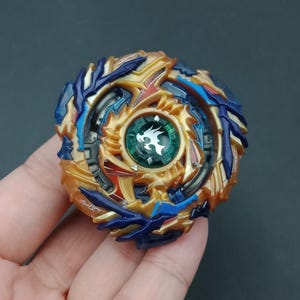 May include: A colorful spinning top toy with a gold, blue, and red design. The center features a green gem with a white flame symbol. The toy is held in a hand against a dark background.