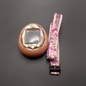 Op de afbeelding: Een bruin Tamagotchi digitaal huisdier met een scherm en drie knoppen. Het heeft een roze en paarse band met het woord "Tamagotchi" erop.